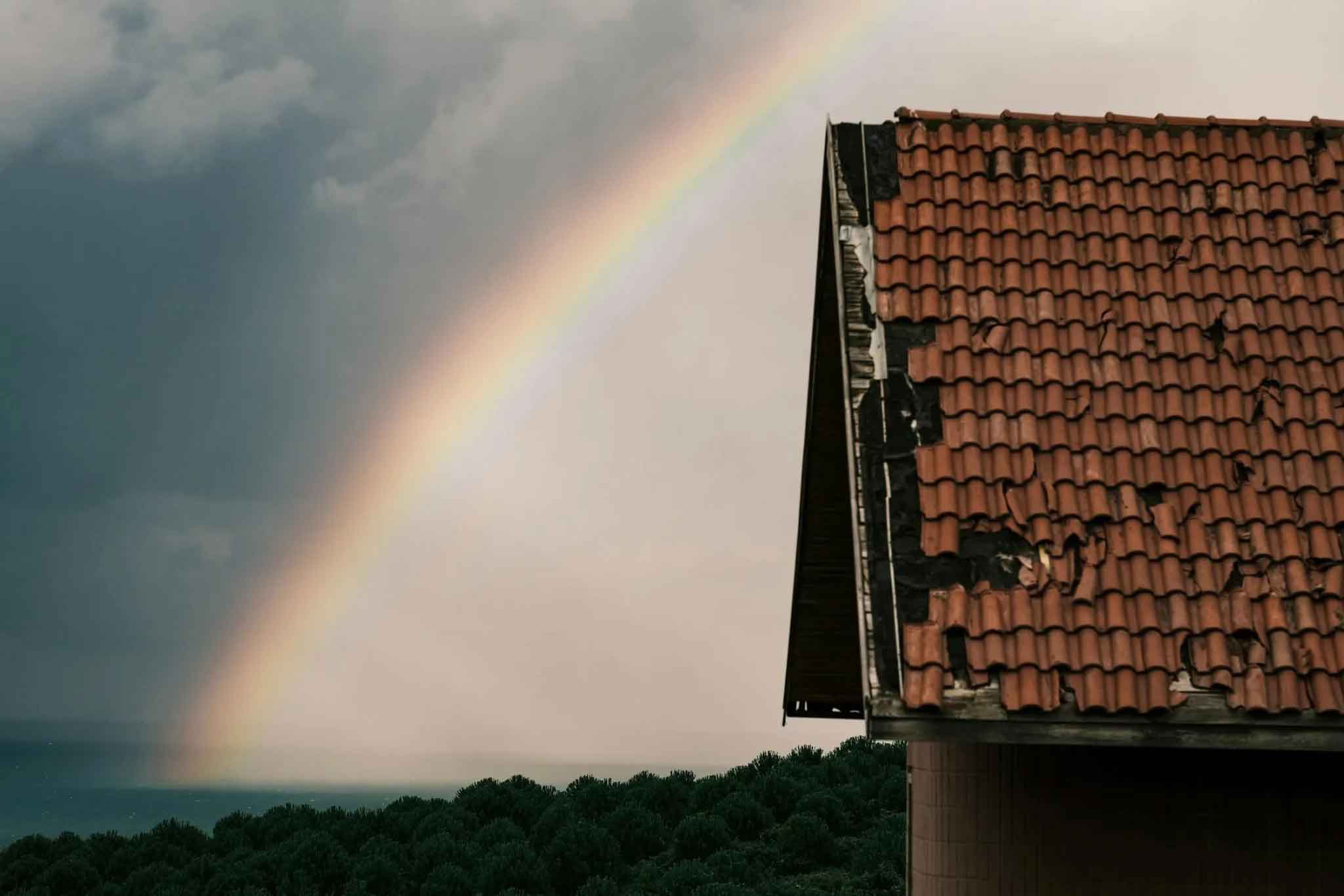 Rainbow in the sky beside a house with a damaged red tiled roof